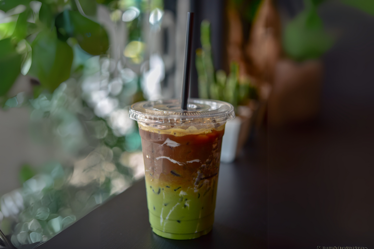 A layered iced dirty matcha latte in a clear plastic cup with a black straw, showing the separation of matcha and espresso over ice, placed on a dark wooden surface with blurred greenery in the background.