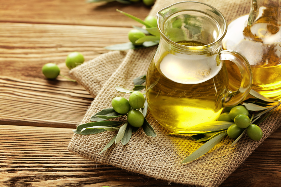 Olive oil placed on a burlap cloth with fresh green olives and olive leaves on a wooden table.