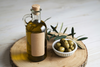 A glass bottle of extra virgin olive oil with a cork stopper, sitting next to a small white bowl of fresh green olives on a wooden round.