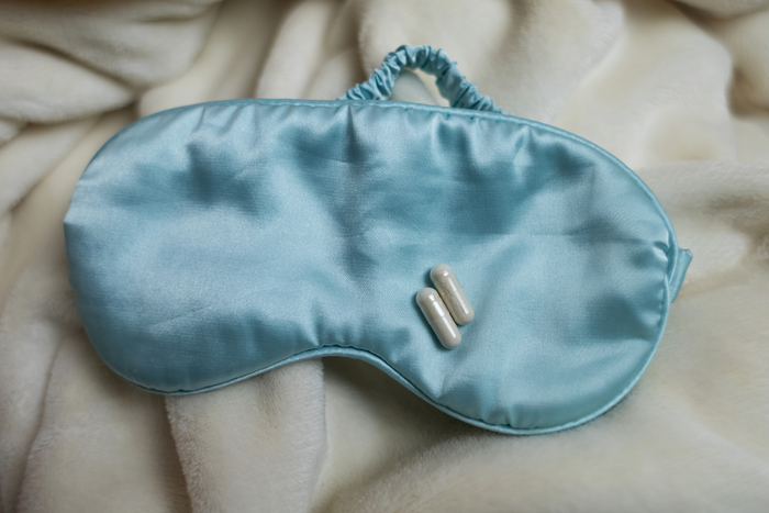 A light blue satin eye mask with two white capsules resting on it lies on a soft, white, fuzzy blanket.
