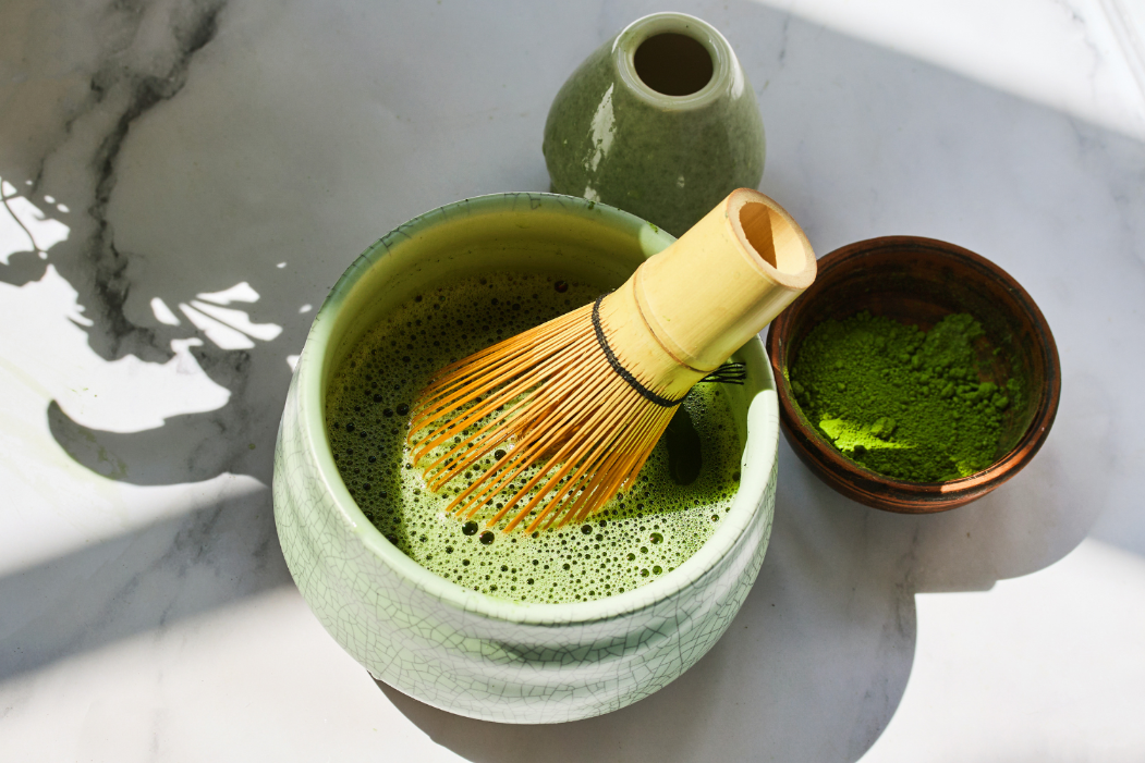 A matcha whisk is in a light green bowl filled with frothy green matcha tea, next to a small bowl of matcha powder and a green vase on a marble surface.