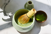 A matcha whisk is in a light green bowl filled with frothy green matcha tea, next to a small bowl of matcha powder and a green vase on a marble surface.