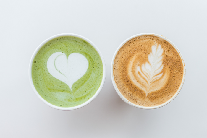 Two cups of latte art placed side by side on a white surface — the left cup contains a vibrant green matcha latte with a heart design, while the right cup holds a brown chai latte with a leaf-shaped design in the foam.