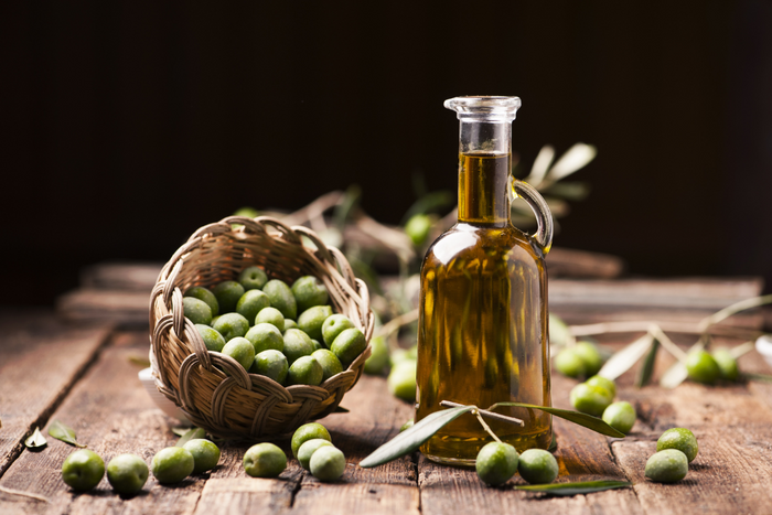 Bottle of olive oil next to a basket of fresh green olives on a rustic wooden table