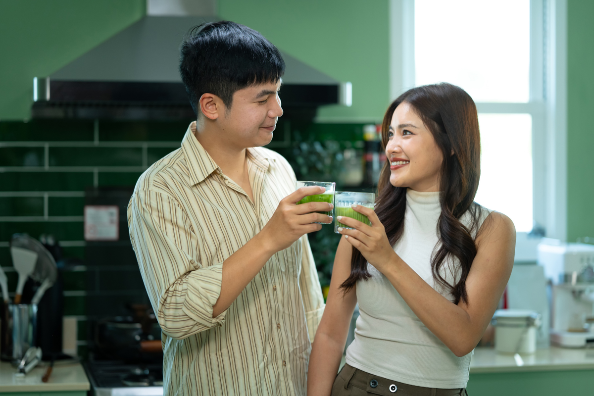 A couple in a kitchen holding up glasses of matcha green tea, smiling at each other.