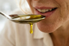 A spoonful of golden liquid, possibly oil or syrup, is held up to a person's mouth, with a drop about to fall.