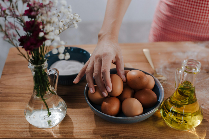 A hand reaches for a bowl of brown eggs, with a vase of flowers, a bowl of sugar, and a bottle of olive oil on a wooden table.