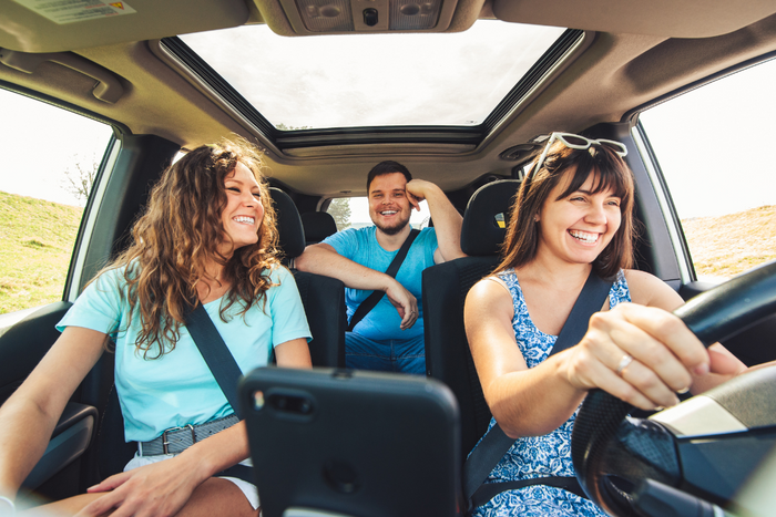 Three friends smiling and talking while driving on a sunny road trip.
