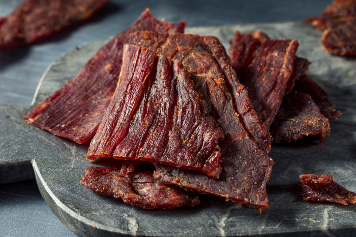 Grass fed beef jerky pieces on a dark stone surface, showcasing rich reddish-brown color, visible meat fibers, and seasoning specks.