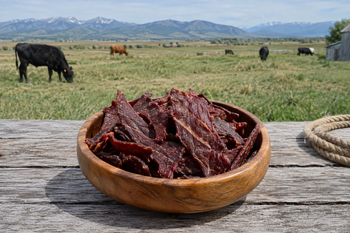 Bowl of grass-fed beef jerky on a rustic table with grazing cattle in the background.