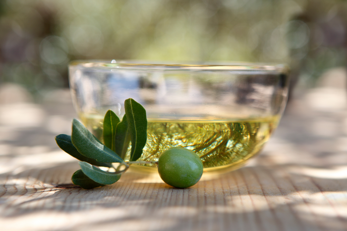 Clear glass bowl of high polyphenol olive oil with green olive and fresh olive leaf on wooden table