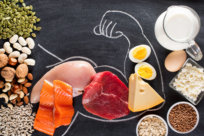 Assorted protein-rich foods including meats, eggs, nuts, and seeds arranged on a chalkboard with a muscle drawing
