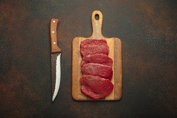 Lean raw beef slices on a wooden cutting board beside a knife for a best beef jerky cuts guide.