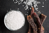 Bowl of coarse salt next to beef jerky strips on a dark surface, representing the search for low sodium beef jerky and lower-salt snack options.