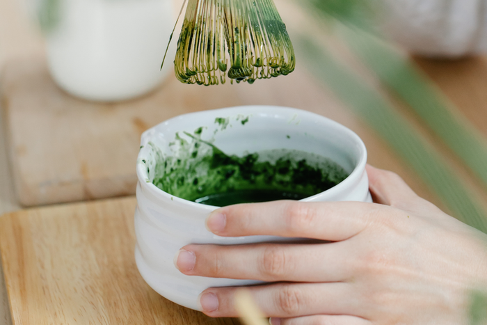A bamboo whisk is used to mix matcha powder and water in a white bowl.