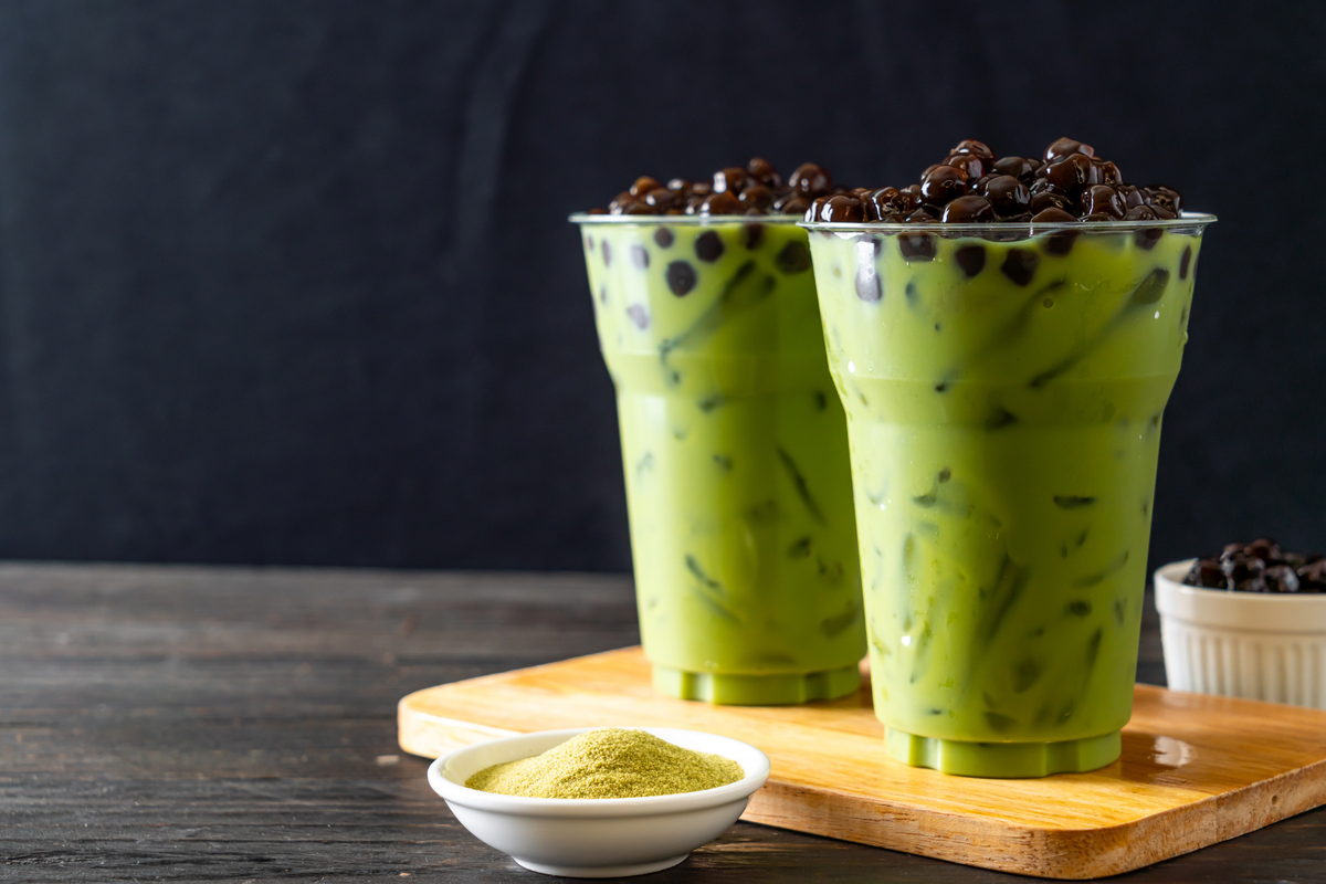 Two tall plastic cups filled with creamy green matcha bubble tea with tapioca pearls, a small bowl of matcha powder, and a bowl of tapioca pearls on a wooden board against a dark background.