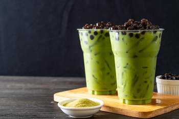 Two tall plastic cups filled with creamy green matcha bubble tea with tapioca pearls, a small bowl of matcha powder, and a bowl of tapioca pearls on a wooden board against a dark background.