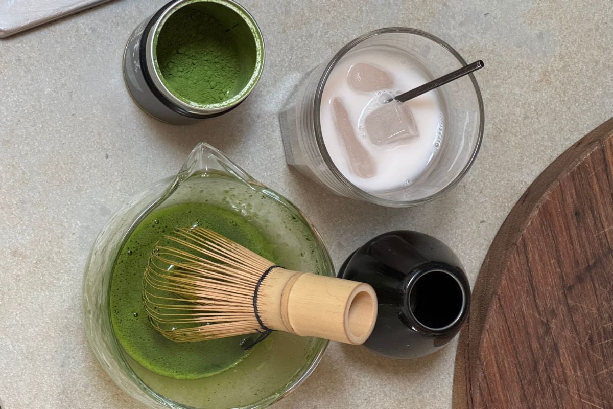 Overhead view of green matcha powder in a tin, whisked matcha with a bamboo whisk in a glass jug, and an iced matcha latte.