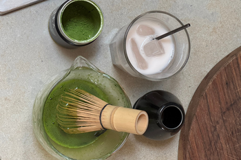 Overhead view of green matcha powder in a tin, whisked matcha with a bamboo whisk in a glass jug, and an iced matcha latte.
