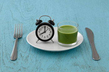 A plate with an alarm clock and a glass of green juice, set on a blue wooden table with a fork and knife.