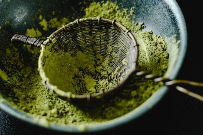 A small metal sieve or sifter resting in a dark ceramic bowl, full of finely ground green matcha powder. Sifting helps prevent clumps and is a step in preparing fresh matcha.