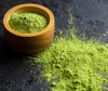 Wooden bowl of vibrant green matcha powder with scattered tea dust showing freshness and calorie details.