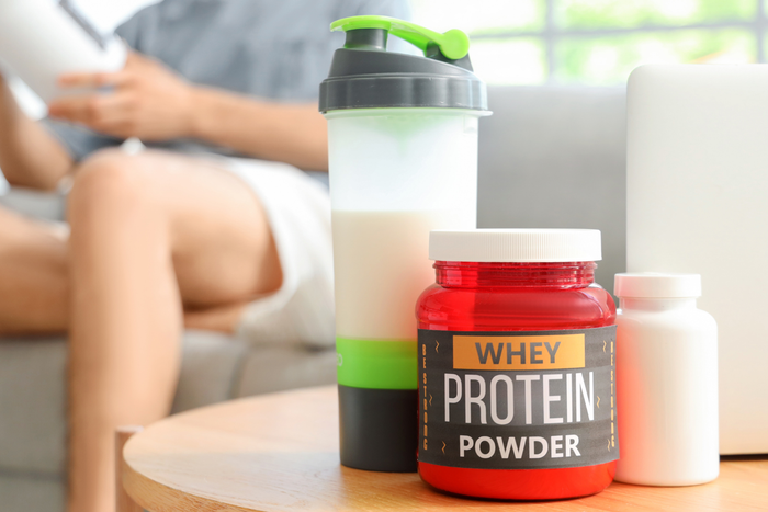 A red jar of whey protein powder, a white bottle, and a shaker bottle filled with a white liquid sit on a table in front of a blurred person.