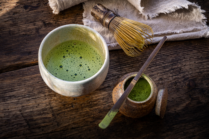 Matcha tea with powder, bamboo whisk, and a wooden spoon beside it.