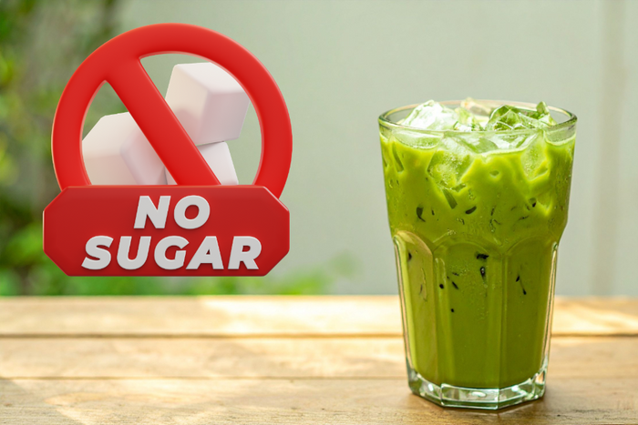 A tall glass of iced matcha latte filled with ice cubes and creamy green liquid beside a no sugar signage