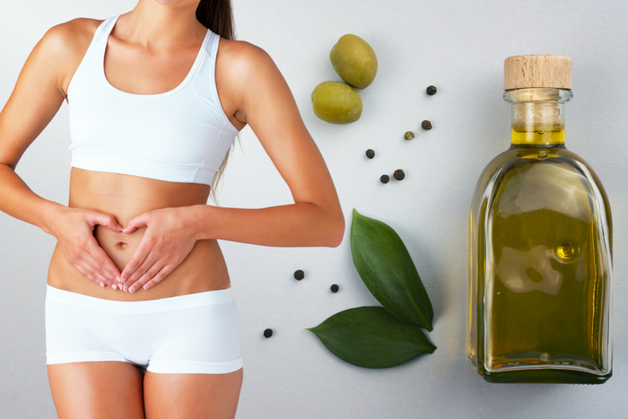 A fit woman making a heart shape over her stomach next to a bottle of olive oil and olives, symbolizing the digestive benefits of EVOO.