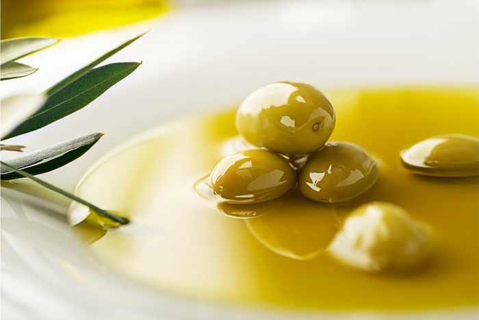 A close-up of green olives resting in a shallow pool of golden olive oil, with an olive leaf on the side for decoration.