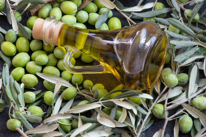 Glass bottle of olive oil resting on a bed of fresh green olives and olive branches.