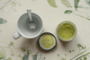 Top view of a ceramic cup with spoon beside a jar of organic matcha green tea powder on a botanical patterned surface