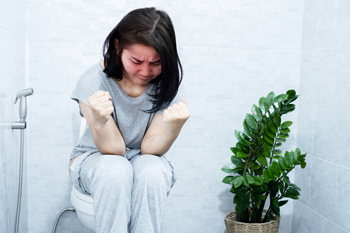 A person sitting on a toilet in a bathroom, wearing grey pajamas.