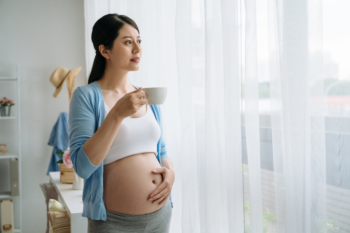 Pregnant woman holding a mug near a bright window while resting one hand on her baby bump.
