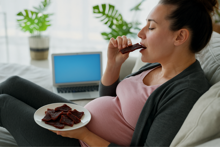 Pregnant woman eating beef jerky on the couch with a plate of jerky