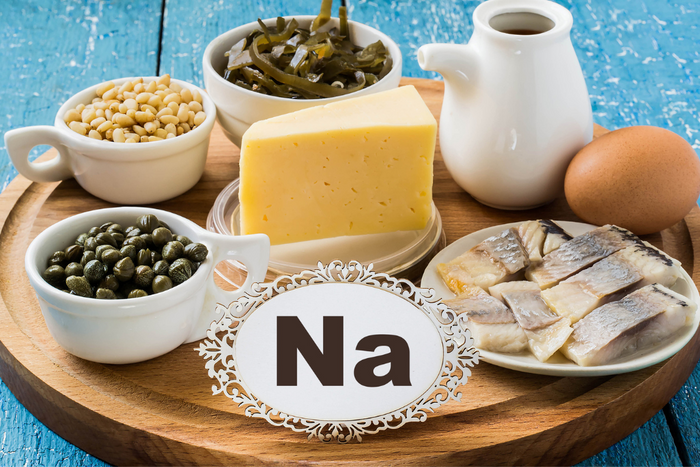 Various sodium-rich foods including cheese, capers, pickled fish, and a sign labeled Na on a wooden tray