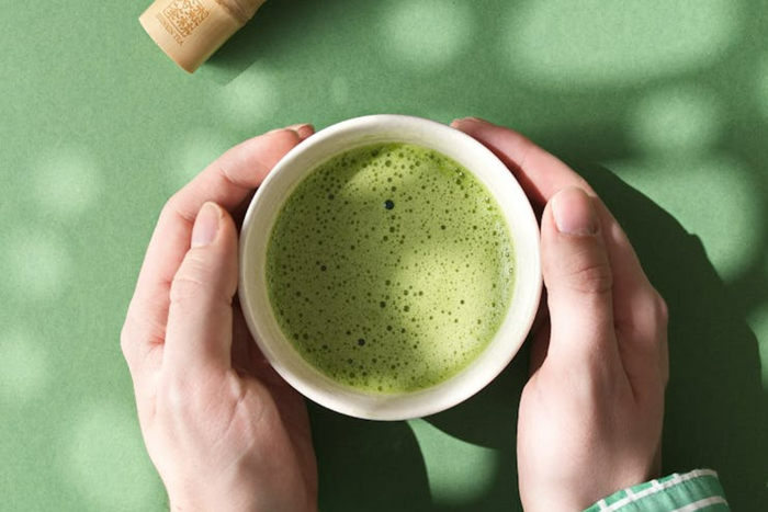 Two hands holding a bowl of freshly whisked warm matcha on green background