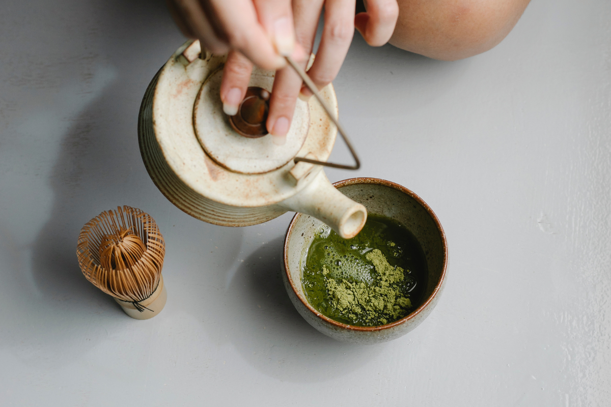 Warm water being poured into a bowl of matcha powder beside a bamboo whisk to prepare smooth ceremonial matcha.