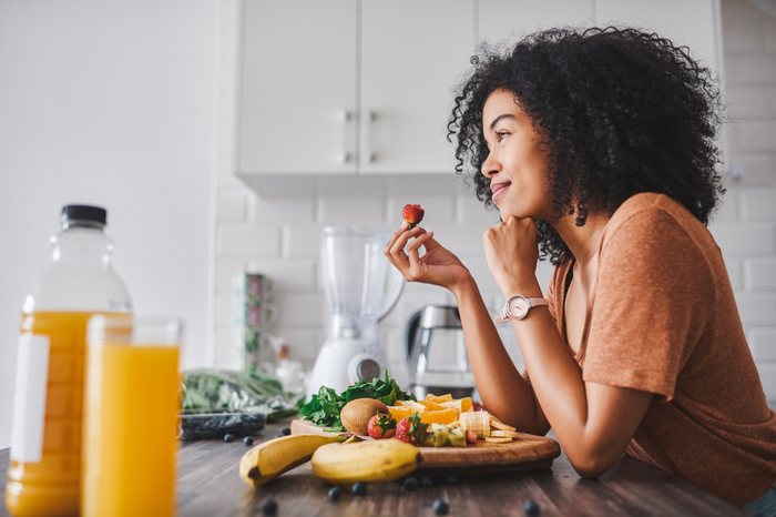 Smiling woman eating fresh fruit at a kitchen table with bananas, oranges, and leafy greens, supporting gut and colon health