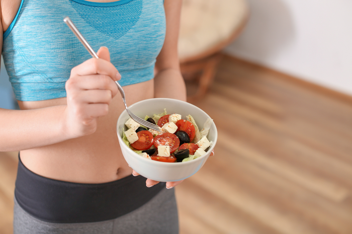Woman in workout clothes eating a fresh salad with tomatoes, olives, and feta after exercise.